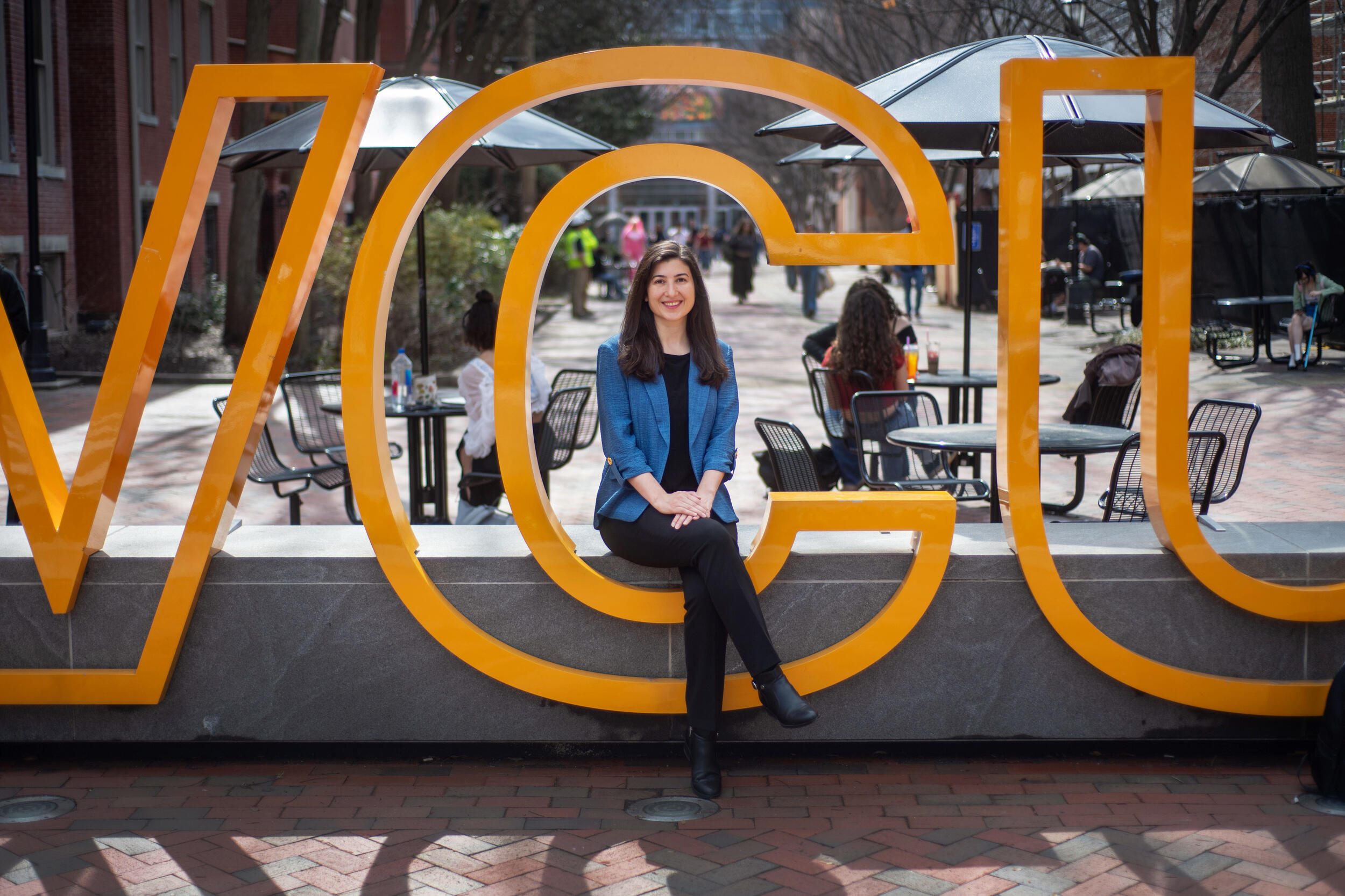 A woman in a blue jacket and black pants sits on a bench surrounded by the C in a large VCU sign.