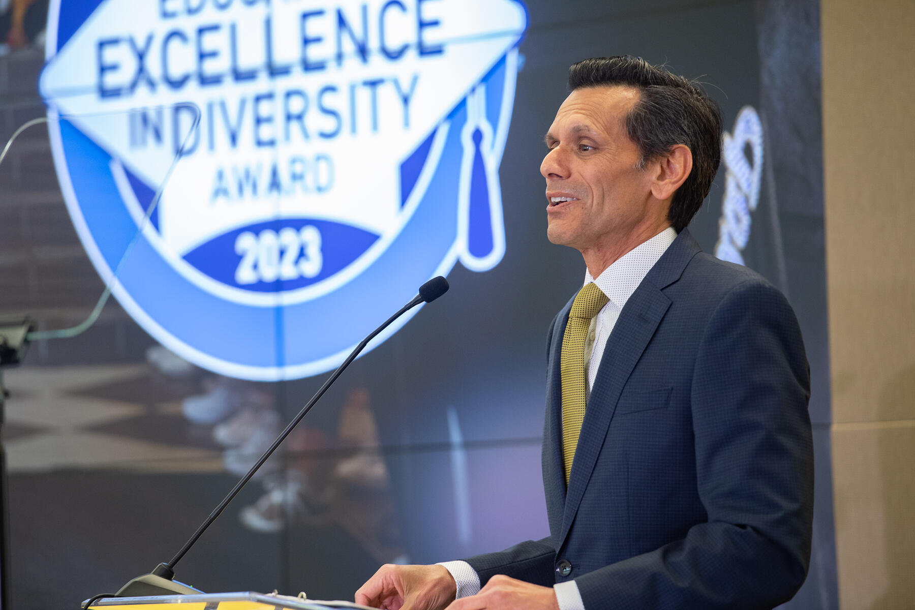 A photo of a man from the waist up. He is facing the left and standing in front of a podium. The man is speaking into a microphone. 