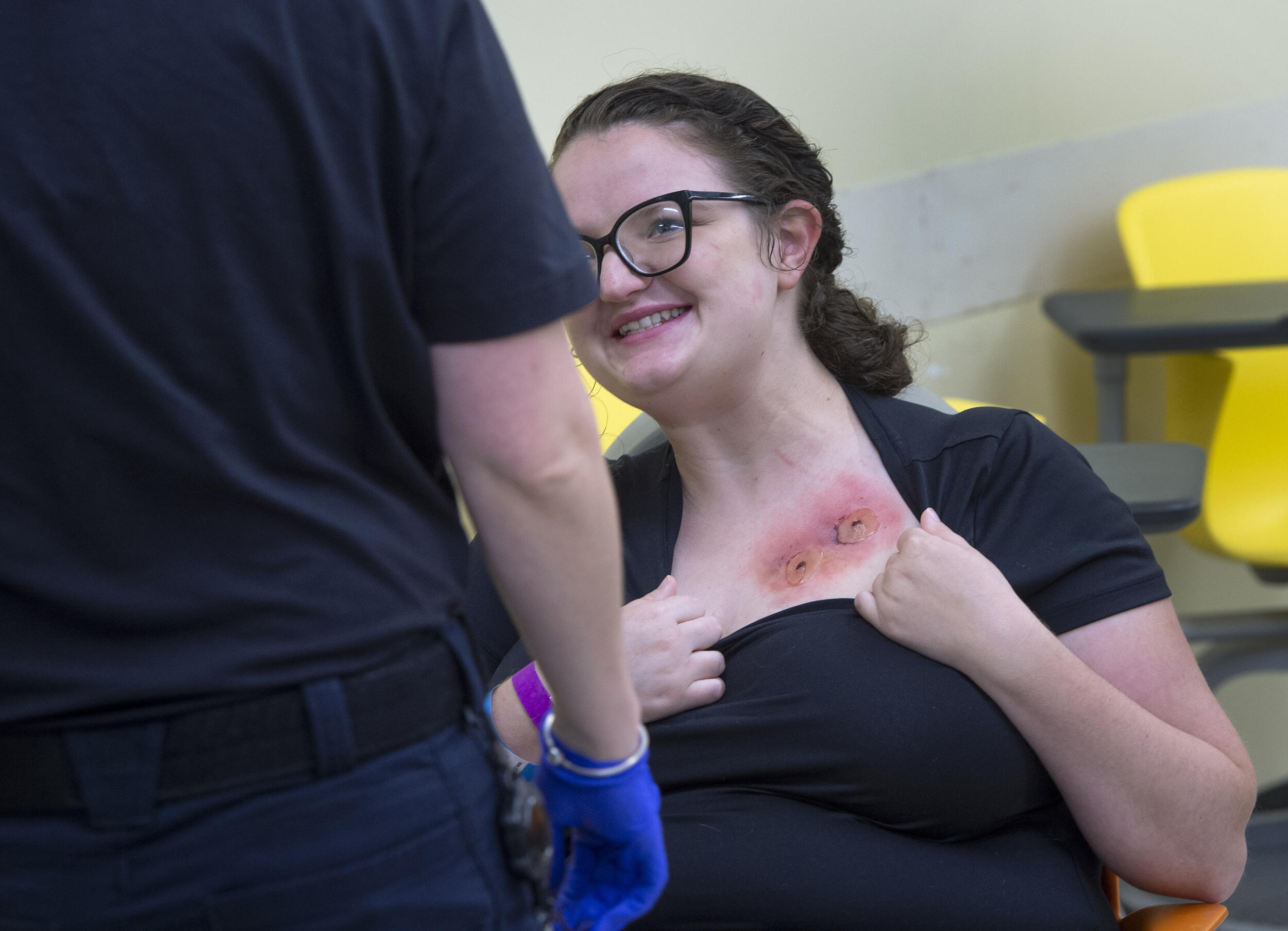A woman smiling while holding down the color of her shirt to show two wounds made with special effects makeup. 