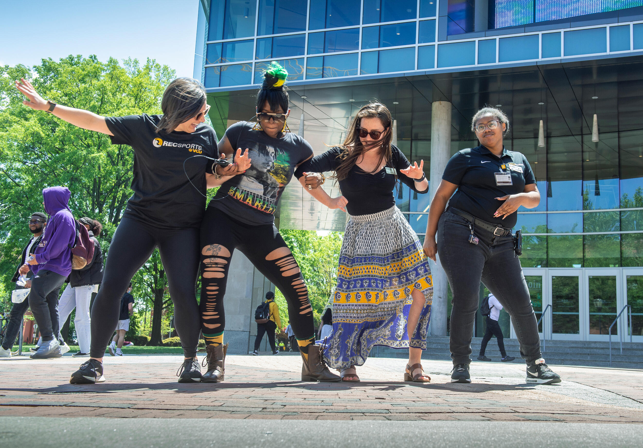 A group of VCU community members dance together in front of the new glass facade of a building on a sunny day on a brick sidewalk.