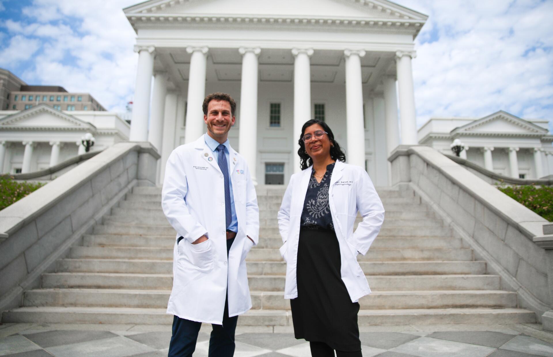 A photo of two people in doctor's coats standing on the steps of the Virginia State Capitol. 