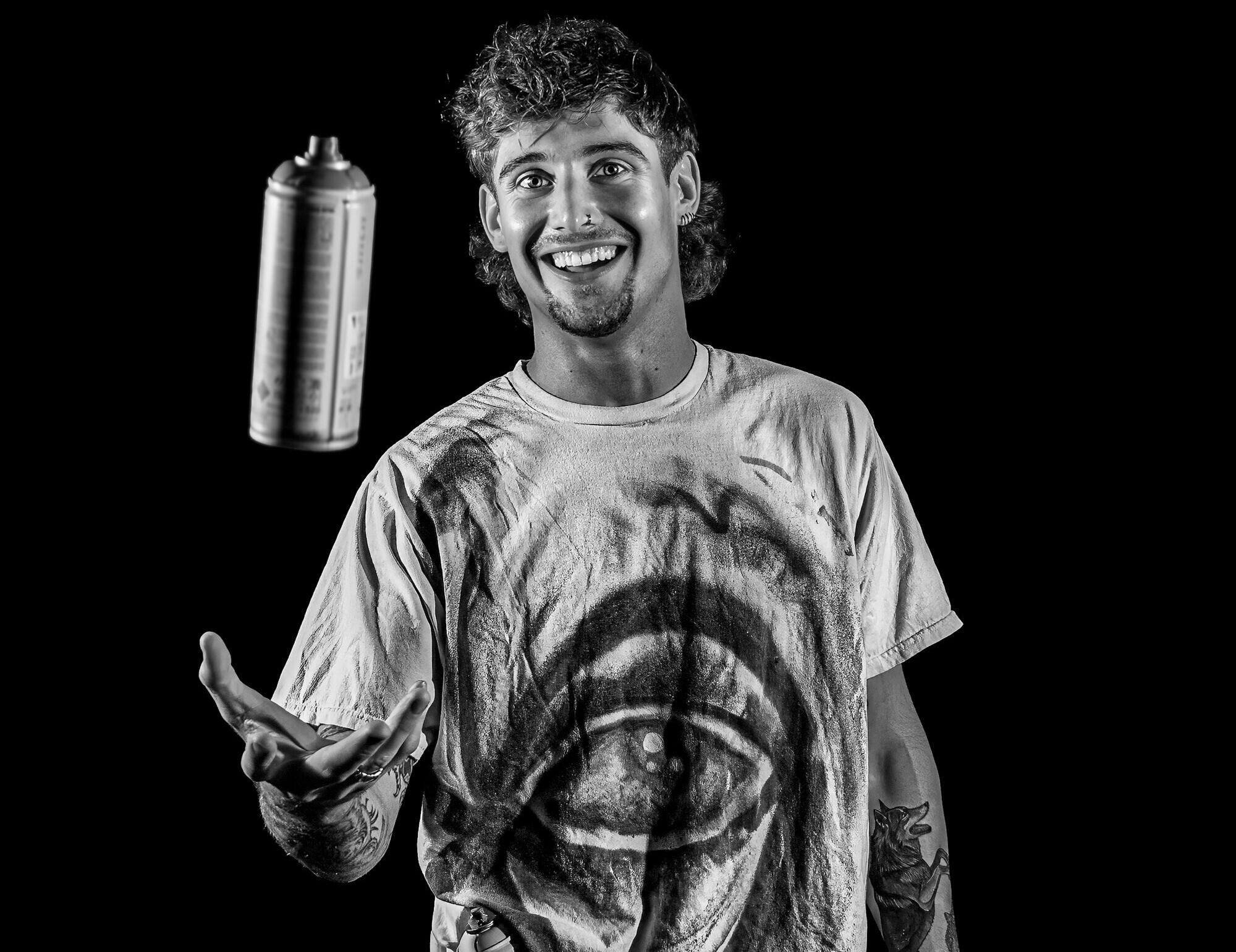 A photo of a man tossing a can of spray paint up in front of him while smiling. 