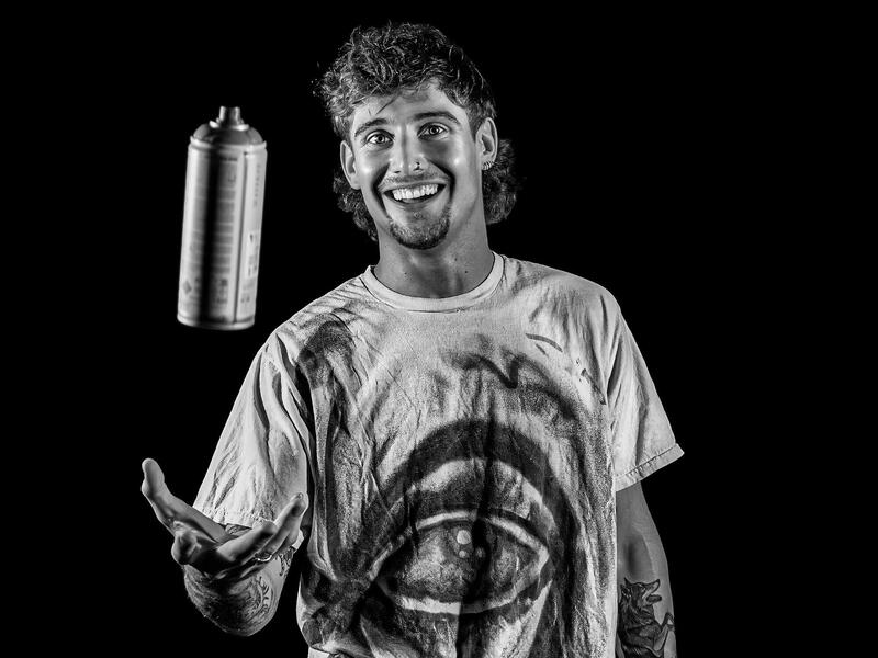 A photo of a man tossing a can of spray paint up in front of him while smiling. 