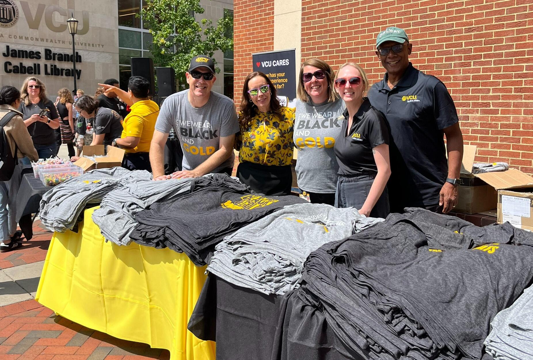 A photo of five people standing behind two tables with yellow and black table cloths. The tables have stacks of T-shirts on them. 