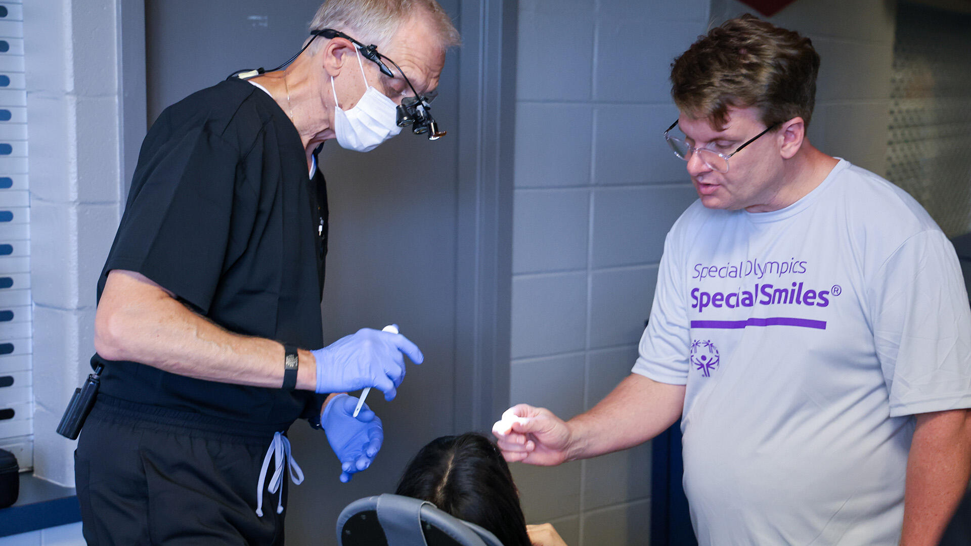 A photo of two men standing over a person in a dental chair. The man on the left is wearing dental scrubs, blue latex gloves, a face mask, and magnifying glasses. The man on the right is wearing glasses and a t-shirt that says \"Special Olympics Special Smiles\" in purple text. 