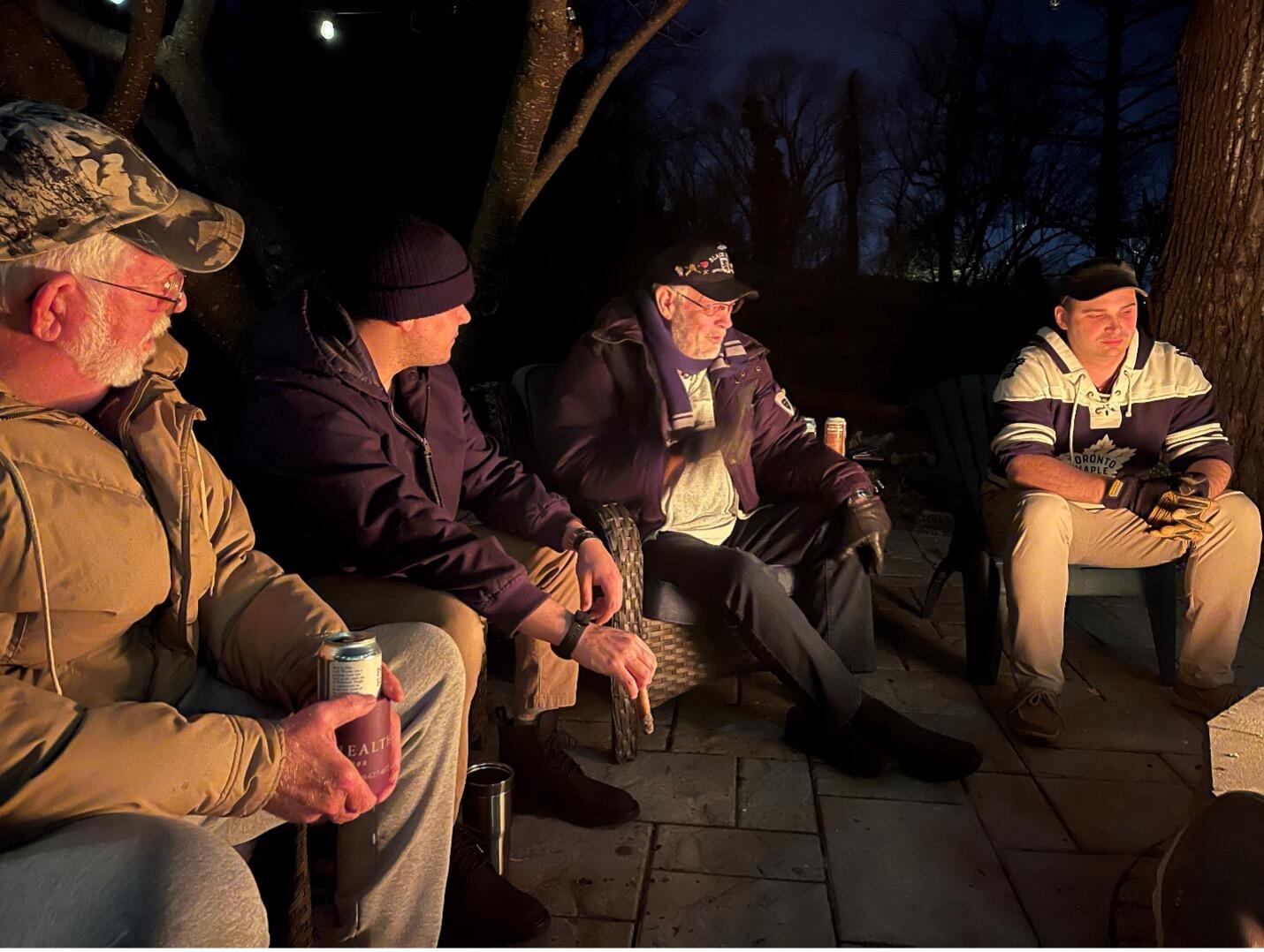 A photo of four men sitting in a semi-circle in the dark. A light from the center of the circle is illuminating them.
