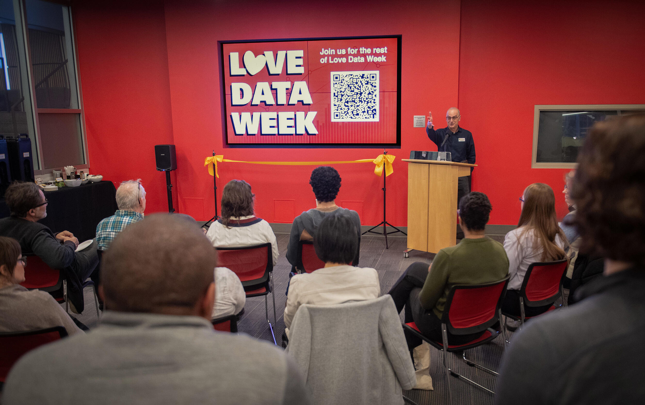 A photo of a room of people looking at a wall with a sign that says \"LOVE DATA WEEK\" in white letters. To the right of the sign is a man standing behind a wood podium and speaking. 