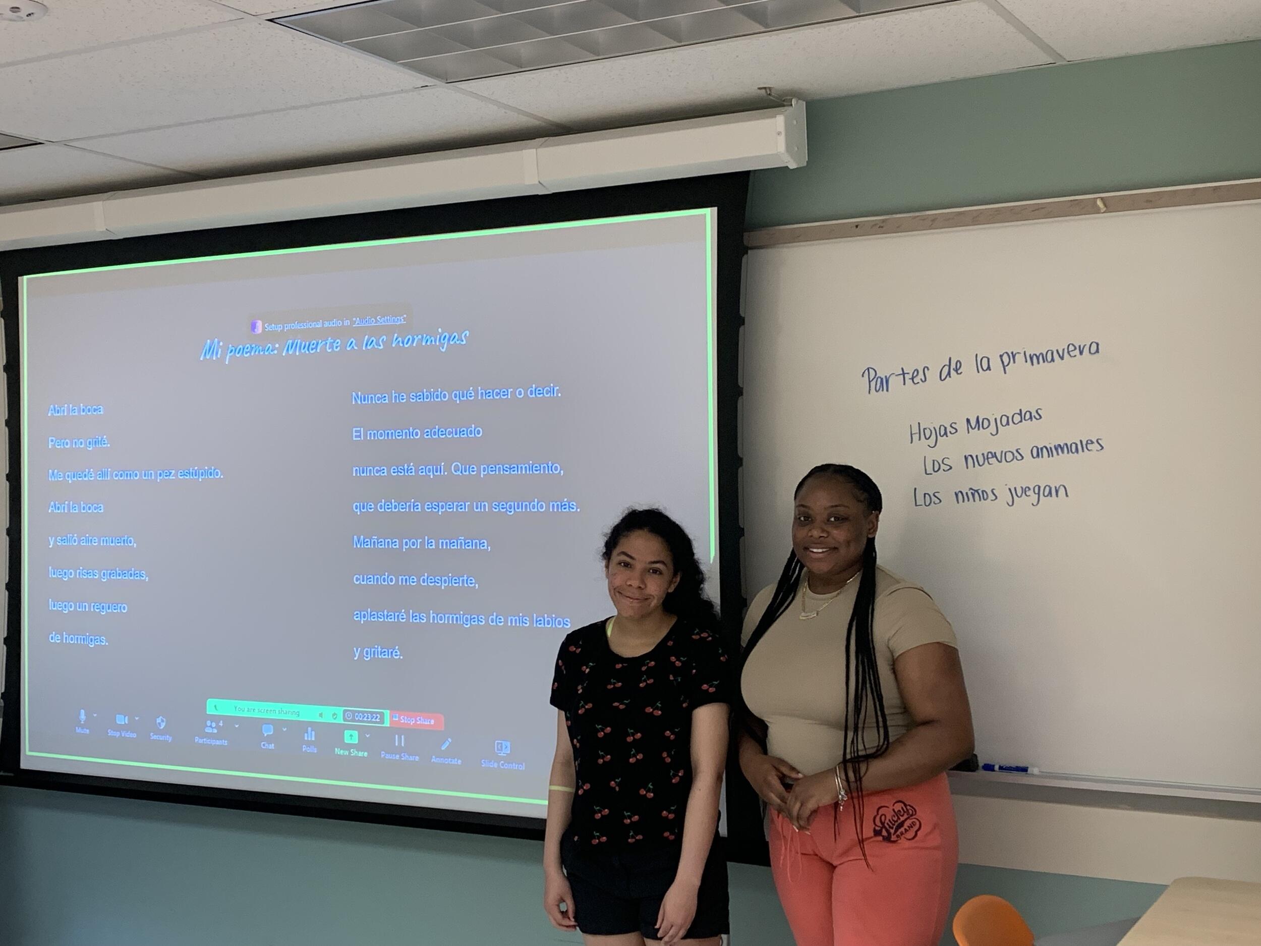 Two women standing in front of a white board and projector screen. 