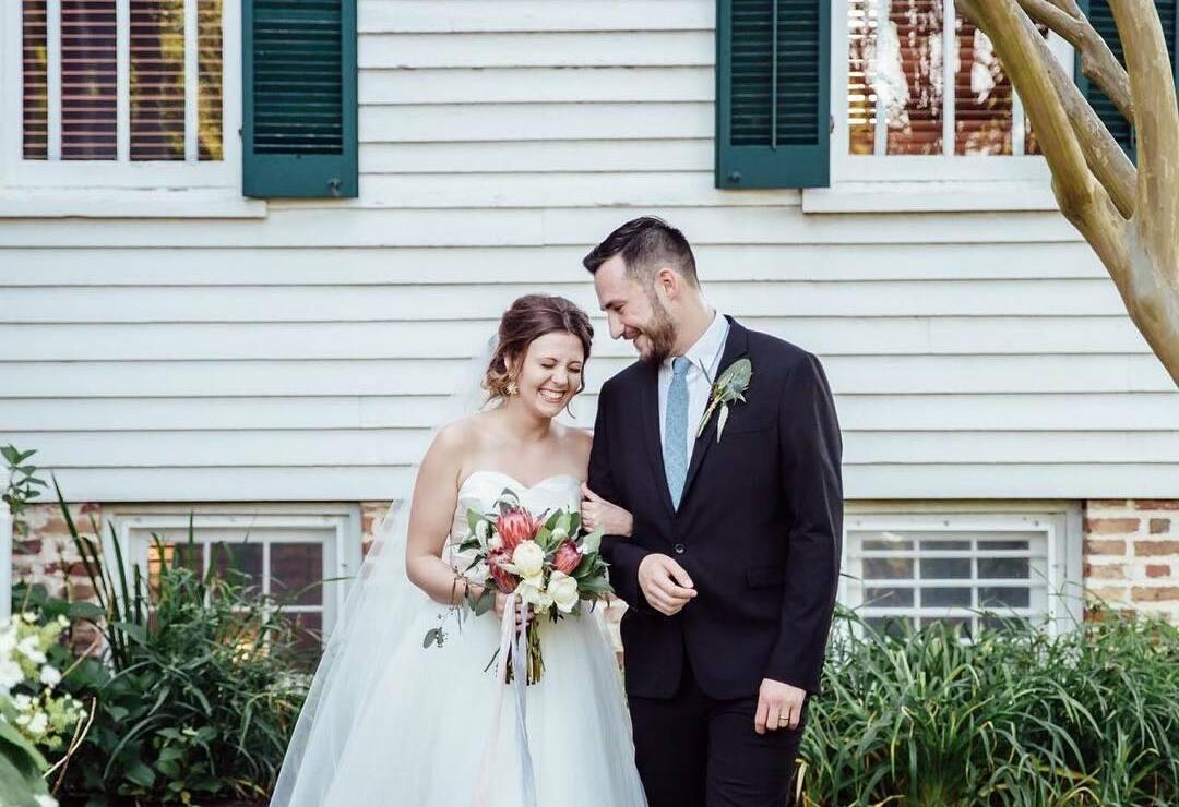 A photo of a woman wearing a wedding dress standing next to a man in a suit and tie. Her right hand is around the man's left arm, and her left hand is holding a wedding bouquet. They are standing in front of a white house with green shutters. 