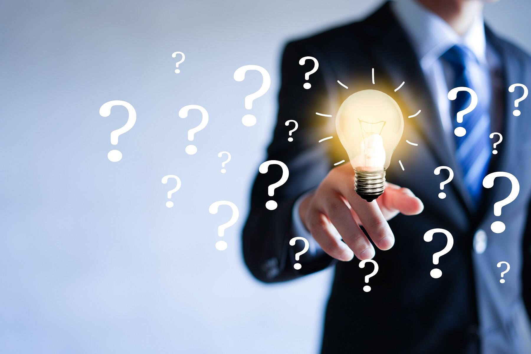 A person in business attire points at a lit light bulb surrounded by question marks.