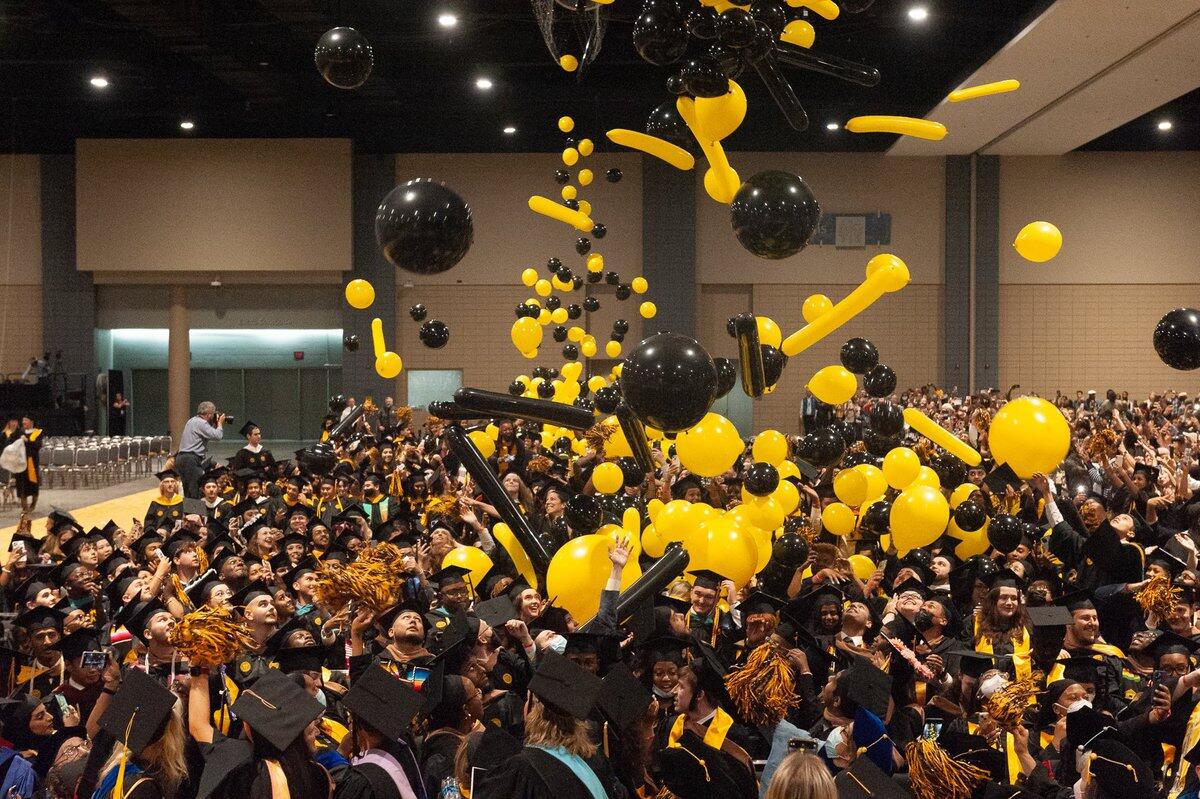 A crowd of graduates wearing graduation caps and gowns with red and yellow balloons falling down on them. 