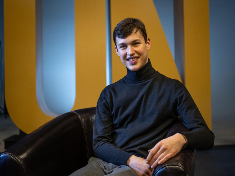 A photo of a man sitting in front of a sign that says \"UN\" in large yellow letters. 