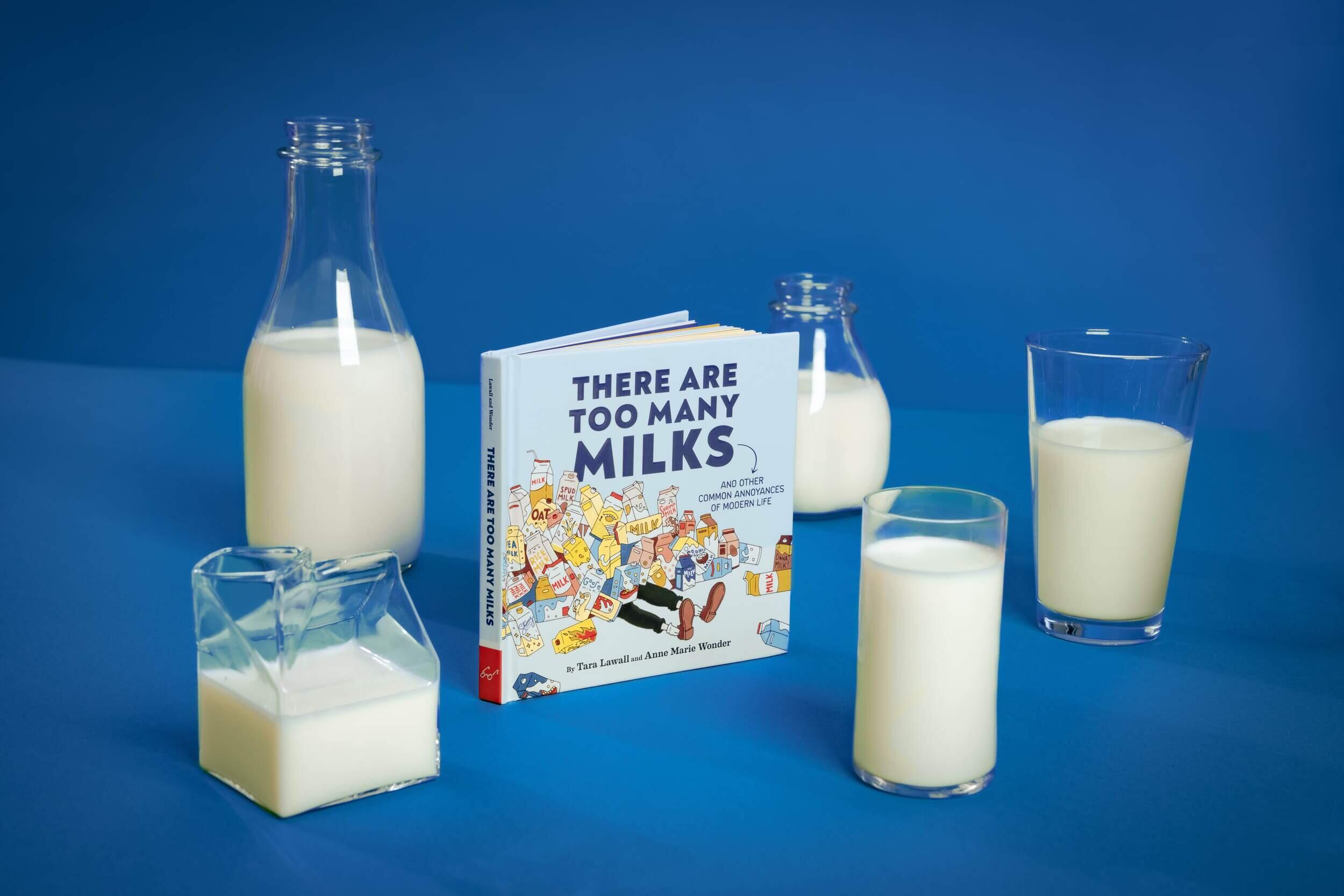 Five glass containers of milk positioned around a book with the title \"THERE ARE TOO MANY MILKS\"