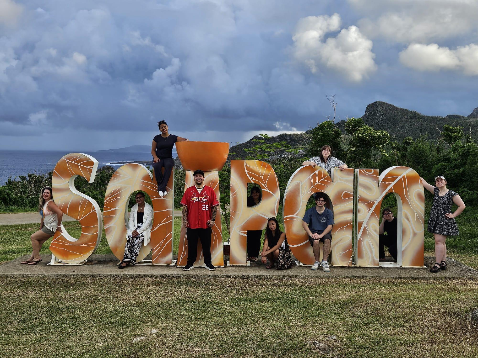 A group of people pose with a large sign spelling out SAIPAN in a field.