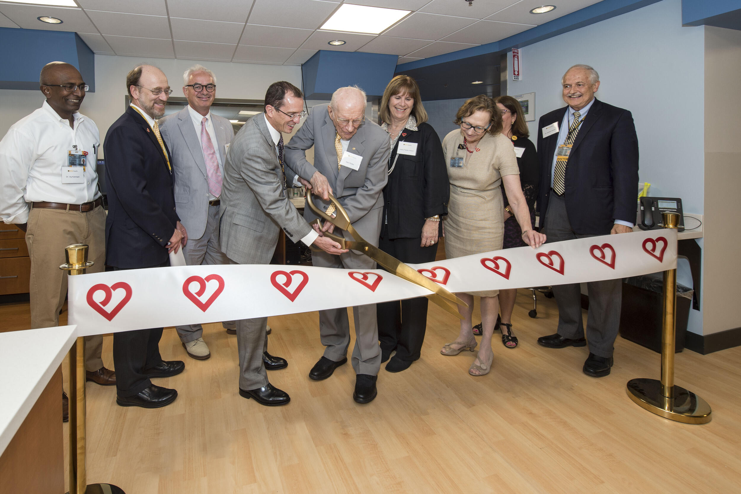 Two people, part of a larger group, use a large pair of scissors to cut a ribbon adorned with the V C U Pauley Heart Center logo.