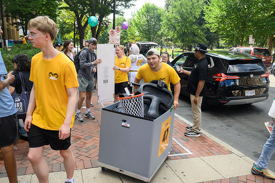 A photo of people moving into a dorm. There is one man pushing a cart with items in it. Around him are people walking and carrying items. Behind him, two people hold onto a floor length mirror. 