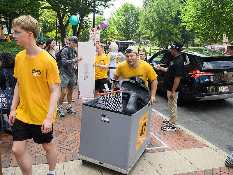A photo of people moving into a dorm. There is one man pushing a cart with items in it. Around him are people walking and carrying items. Behind him, two people hold onto a floor length mirror. 