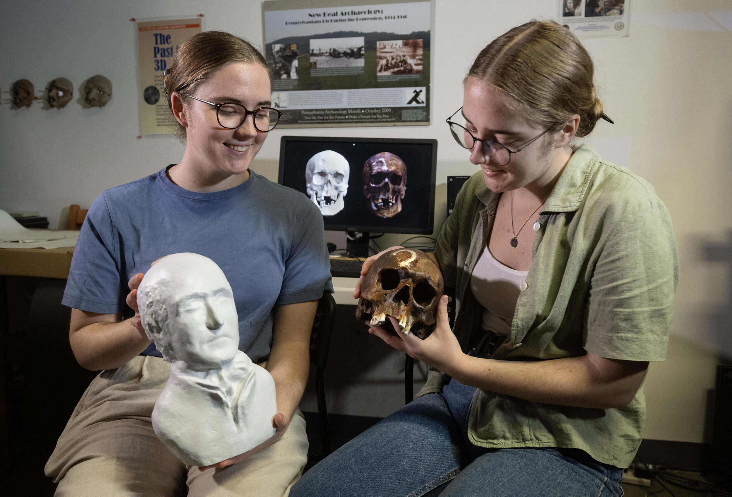 Two women sitting next to each other holding objects. The woman on the left is holding a statue bust, and the woman on the right is holding a skull. 