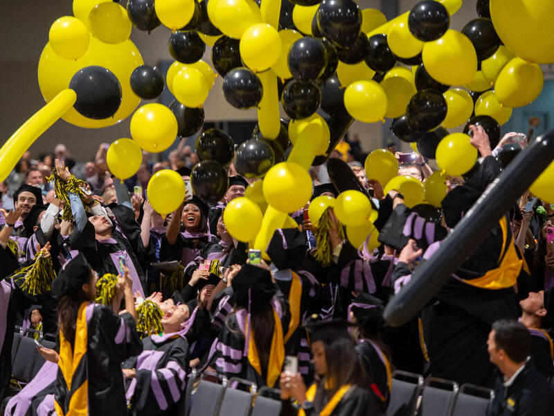 A photo of a crowd of students wearing graduation caps and gowns standing up under a bunch of falling balloons. The balloons are black and yellow.