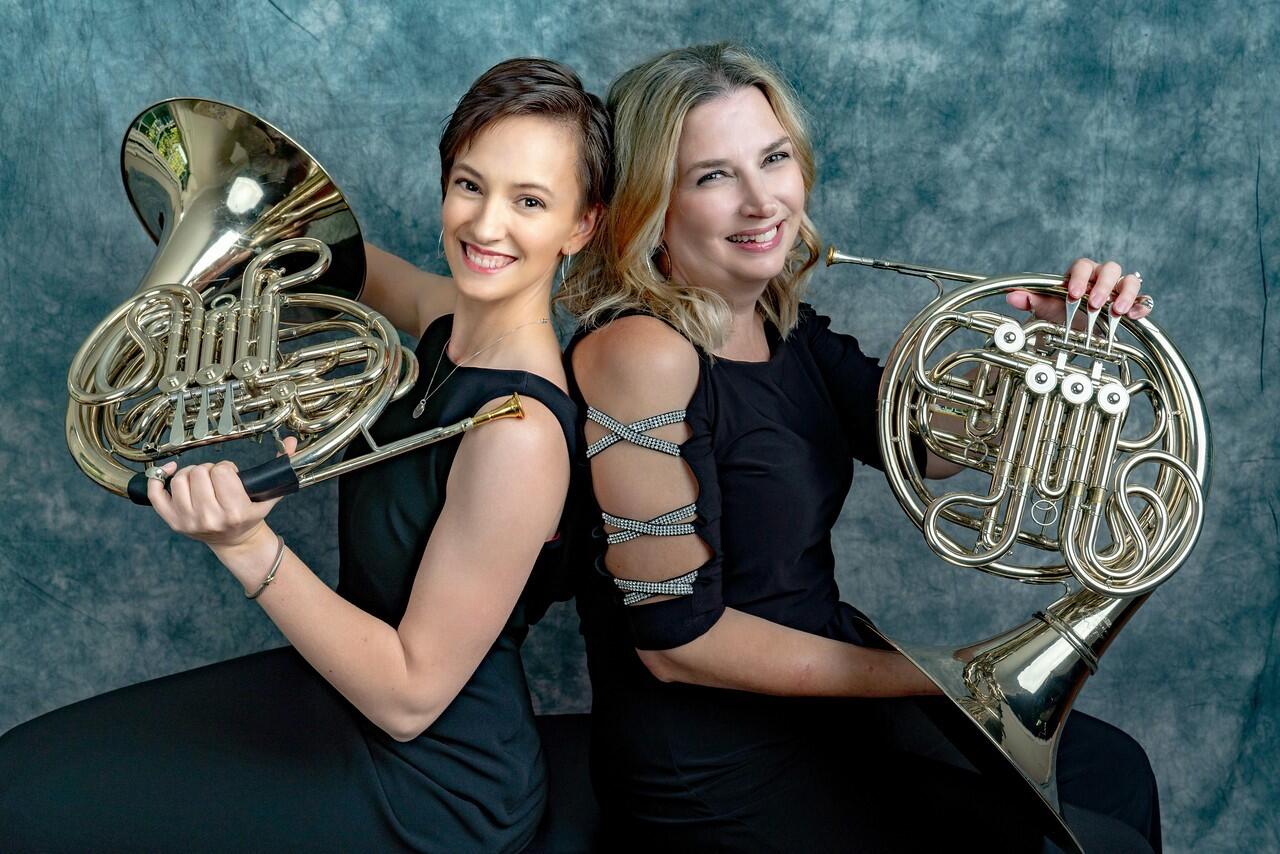 Two orchestra members pose with french horns 