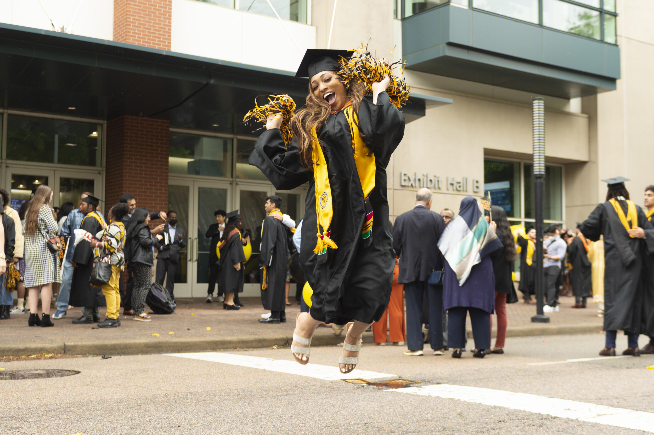 A woman wearing a graduation cap and gown jumping in the air with pom poms 