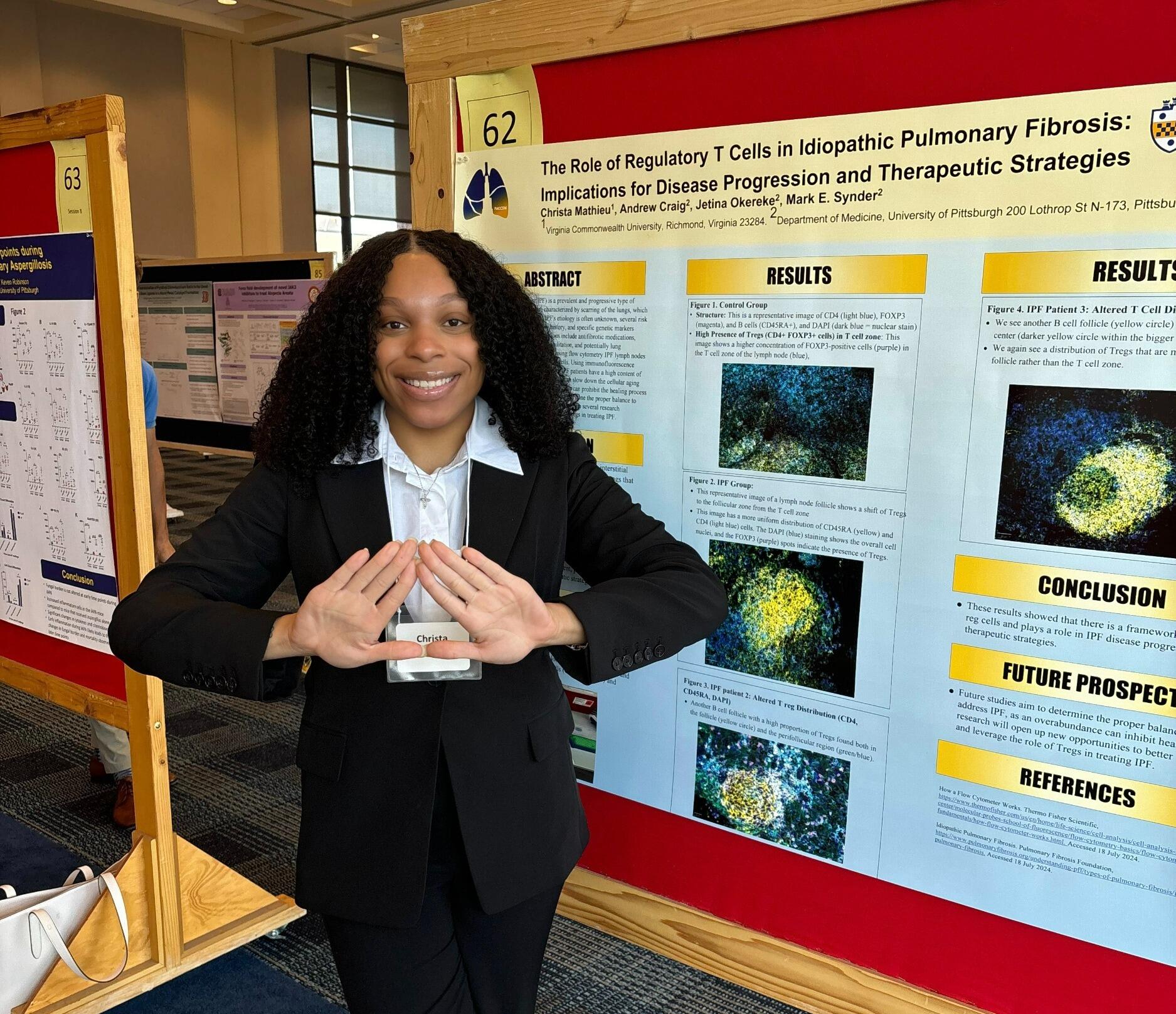 A photo of a woman standing to the right of a research poster. She has her hands in front of her, and they are forming the shape of a heart. 