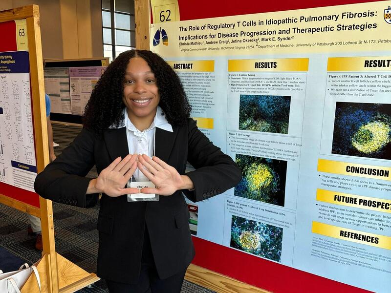 A photo of a woman standing to the right of a research poster. She has her hands in front of her, and they are forming the shape of a heart. 