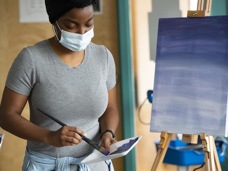 A woman wearing a facemask dipping a paintbrush in blue paint while standing next to a canvas