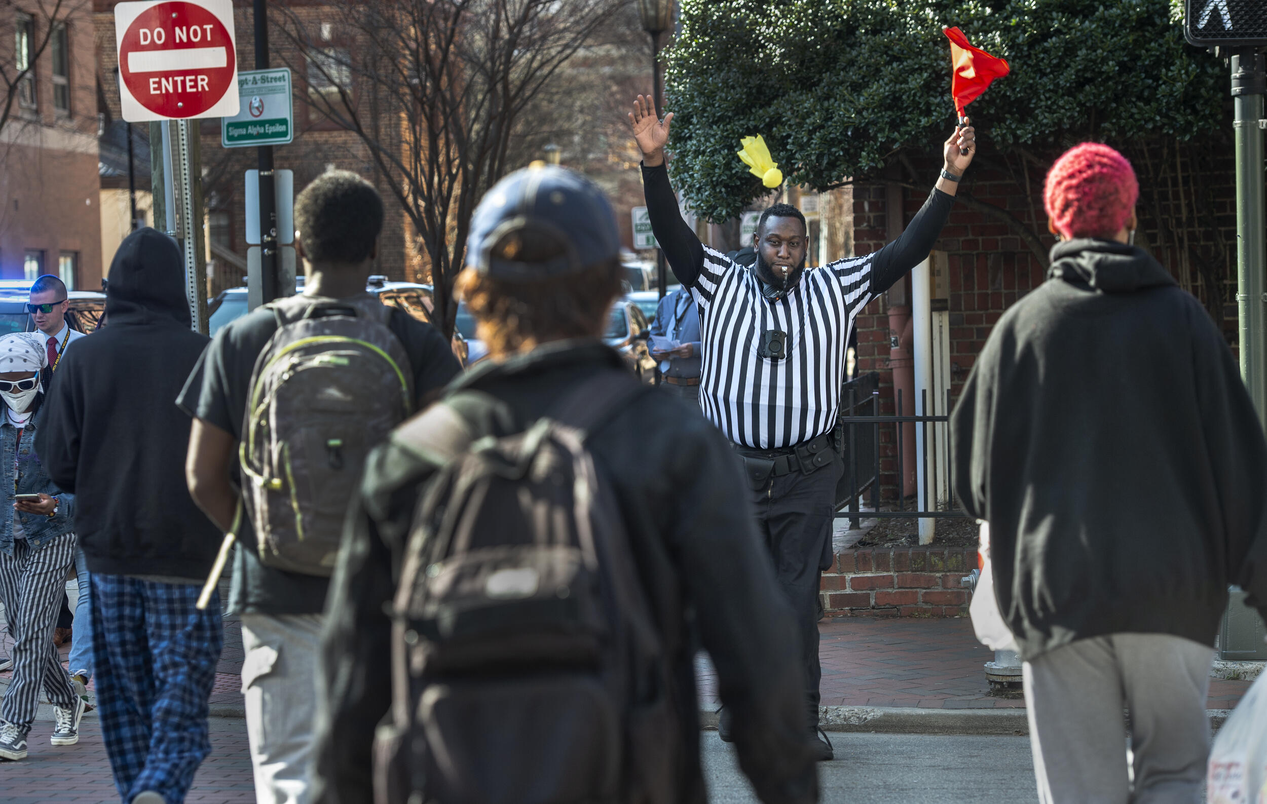 A man in a striped referee shirt blows a whistle and holds up a red flag as people cross the street toward him.