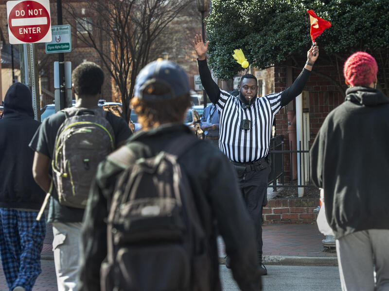 A man in a striped referee shirt blows a whistle and holds up a red flag as people cross the street toward him.
