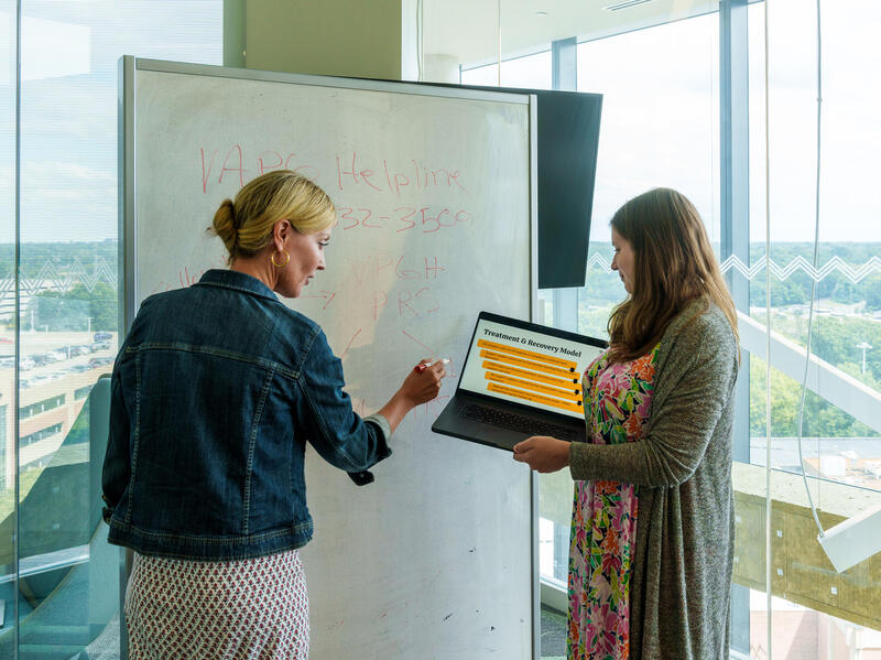A photo of two women standing in front of a white board. The woman on the left is writing on the board and the woman on the right is holding a laptop that they are both looking at. 