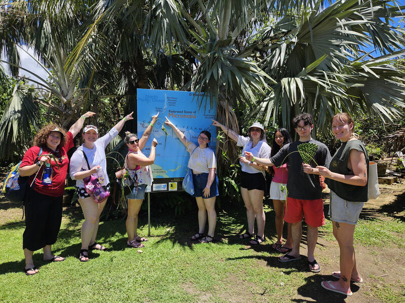 A photo of 8 people pointing at a sign that shows and island on a map. 