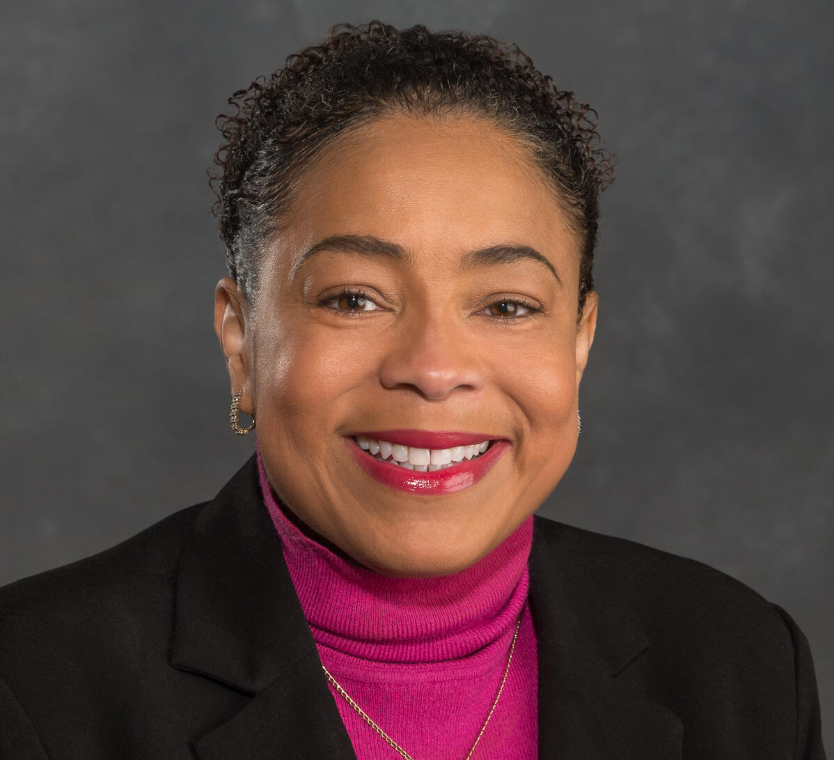 A photo of a woman from the shoulders up. She is wearing a fuchsia turtleneck and a black sports coat. 