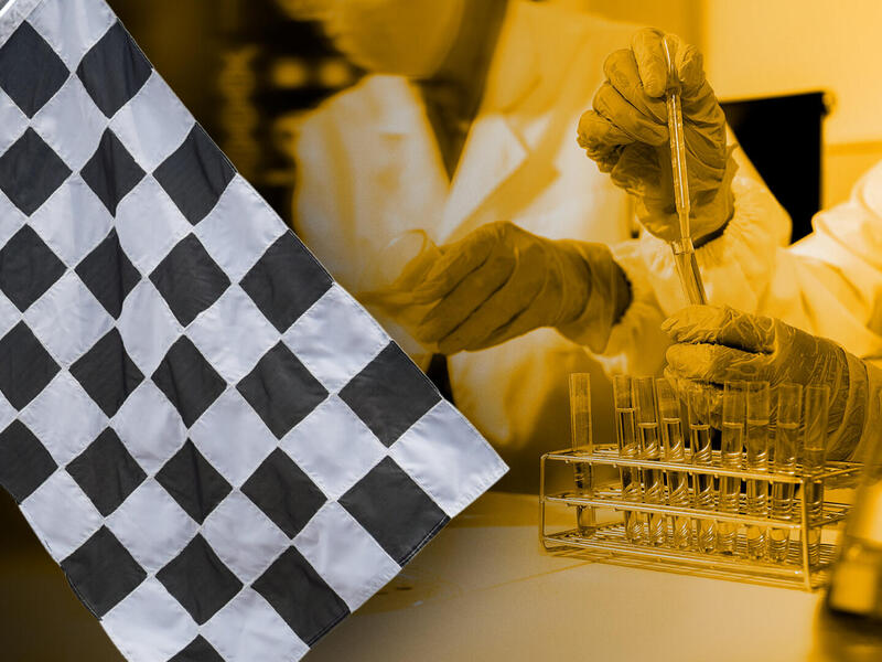 A photo of a black and white checkered flag in front of two researchers who are using droppers to put liquid in beakers. 