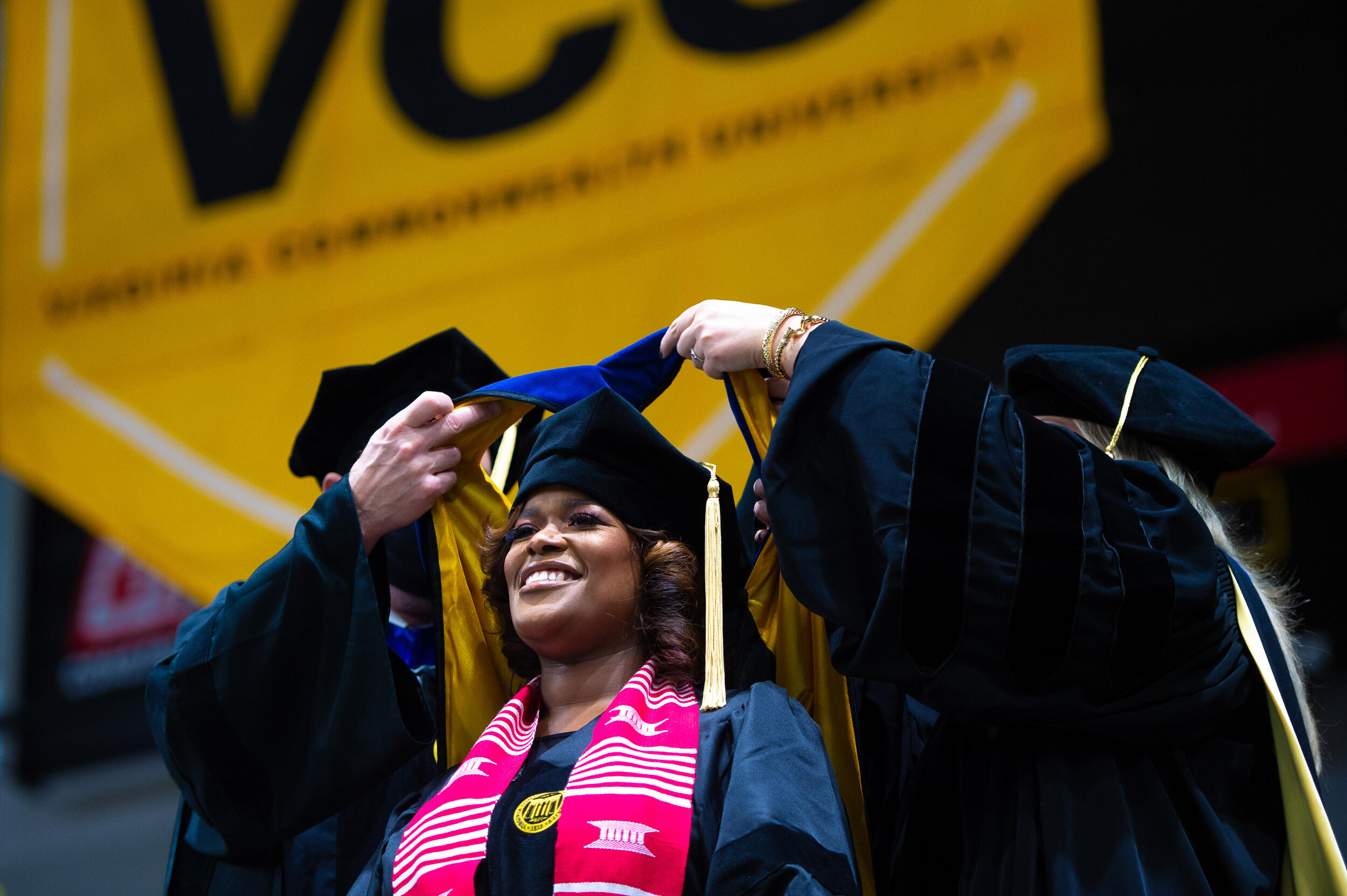 A doctoral student receives a hood at a hooding ceremony, as two professors lift a piece of fabric over their head on the stage in front of a large VCU banner.