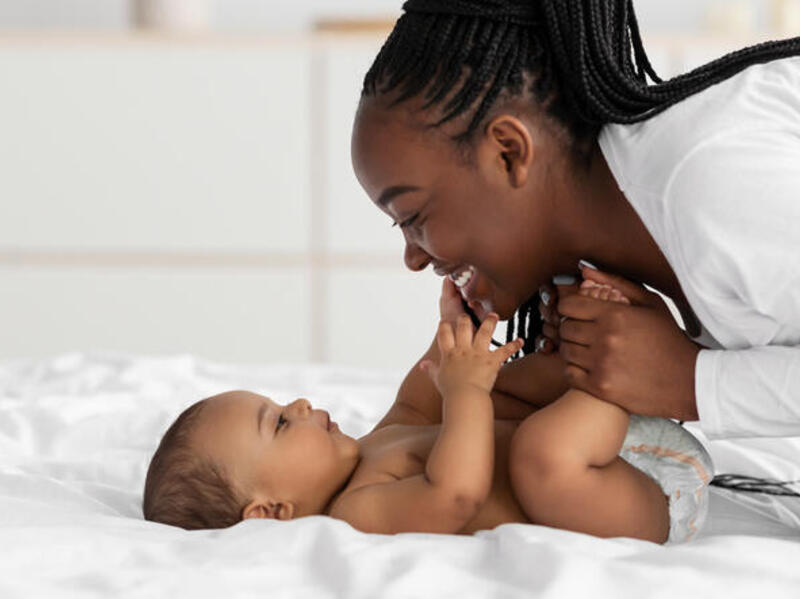 A photo of a woman lenaing over a baby and smilining. She is holding the baby's legs while the baby is lying down, and the baby is touching the woman's face. 