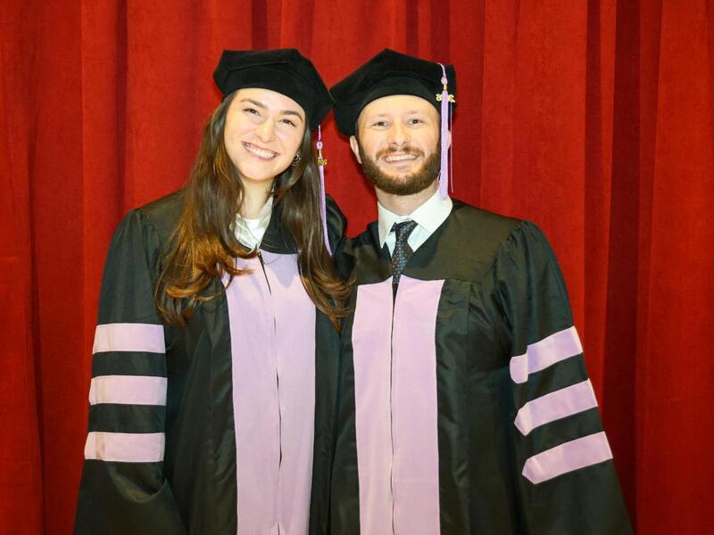 A photo of a man and woman wearing graduation cap and gowns standing next to eachother in front of a red curtain. 