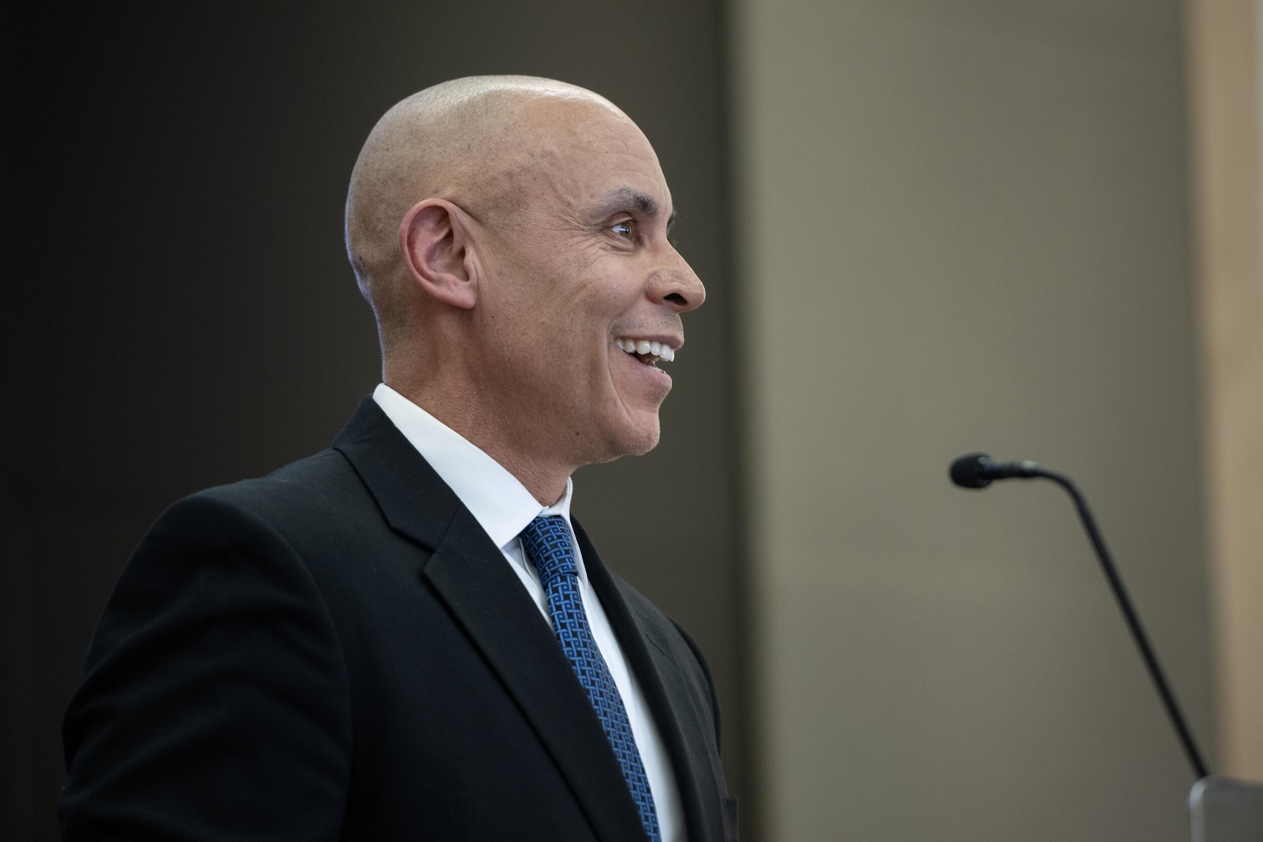 A photo of a man from the chest up. He is wearing a suit and tie and facing the right. In front of him is a microphone. 