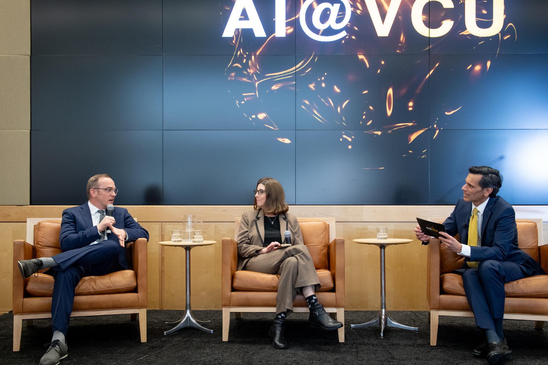 Two men in suits sit on either side of a woman in a suit, all in conversation. A panel in the background reads AI @ VCU