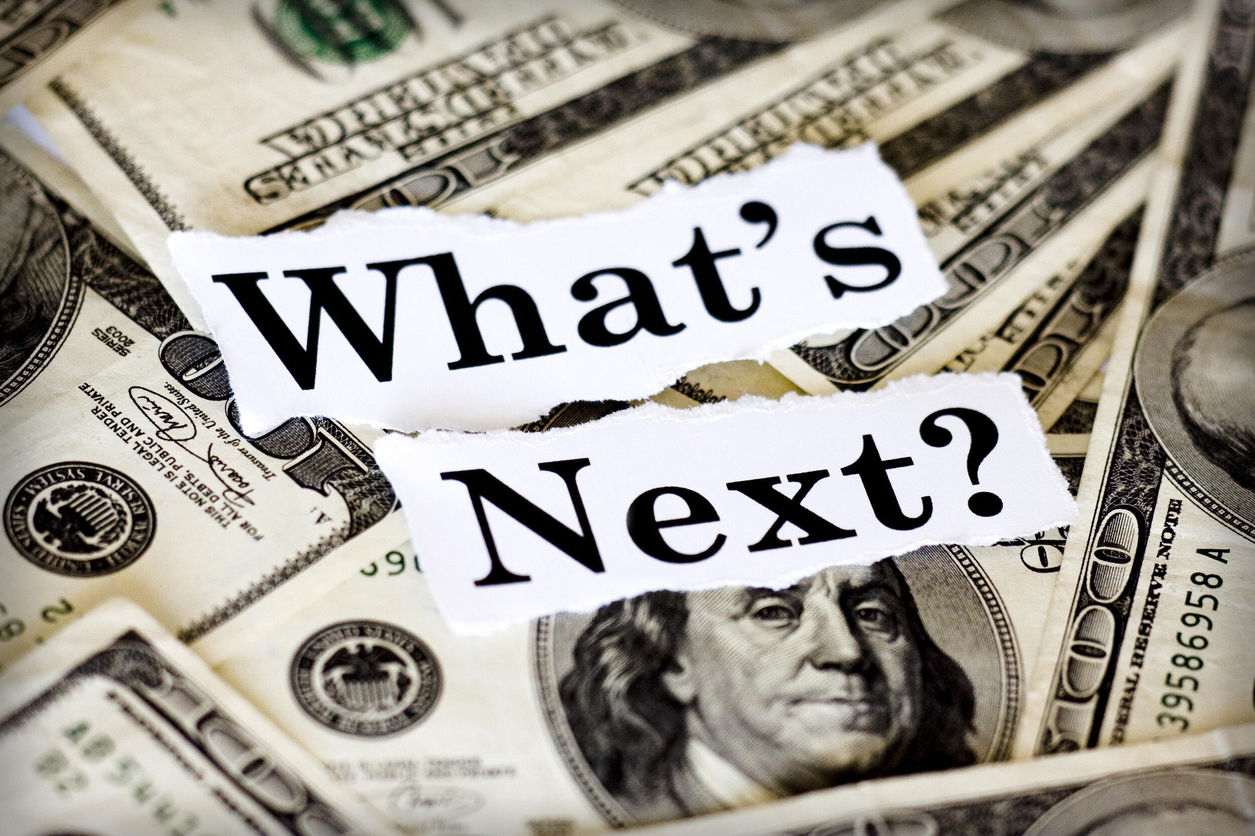 The words \"What's Next?\" in black text on white paper on top of hundred dollar bills 