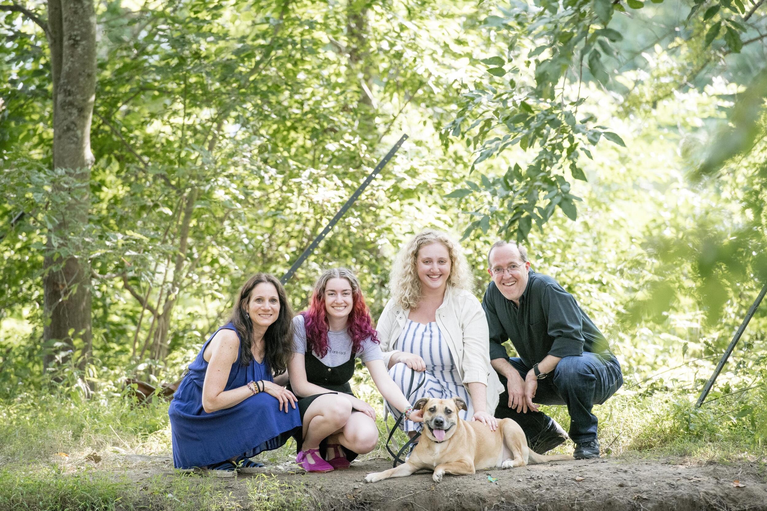 A photo of four people crouching in the woods behind a dog laying on the ground. 