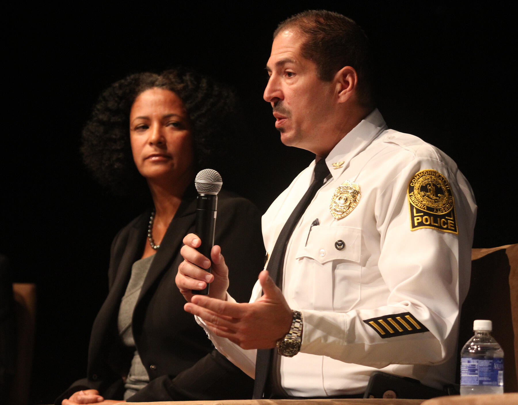 A photo of a man and woman from the waist up. The man is on the right and holding a microphone while speaking. The woman is on the left watching him speak. 