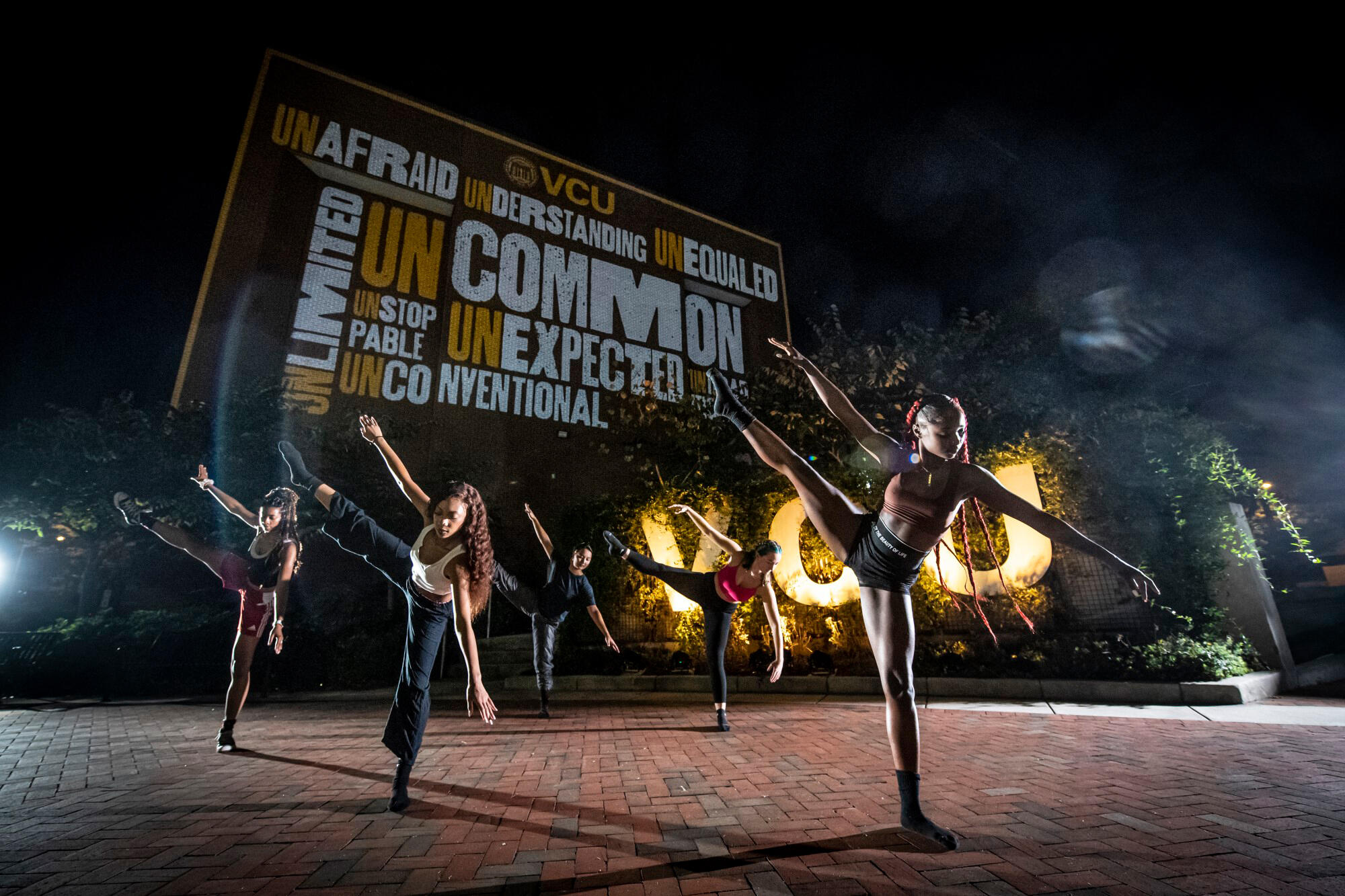 Students participate in a dance performance at night under a wall lit with an artistic word cloud of words that fit with the uncommon experience VCU provides: Unafraid, unlimited, understanding, unequaled, uncommon, unexpected, unstoppable and unconventional.