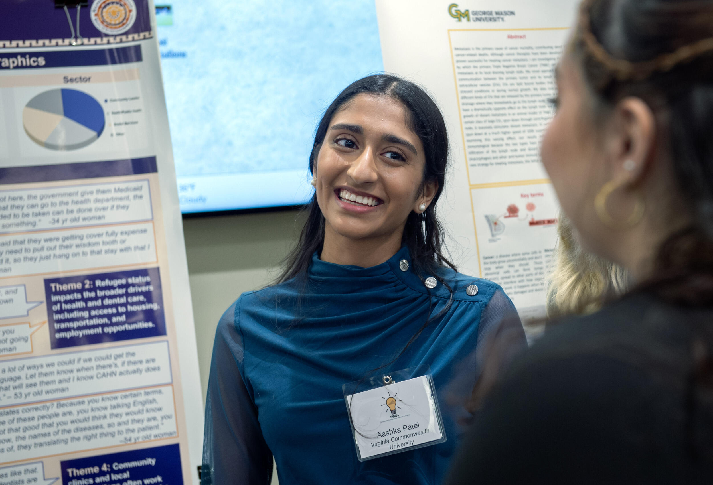 This is a photo of a woman smiling at another woman facing her. The first woman is standing to the right of a poster with research on it. 