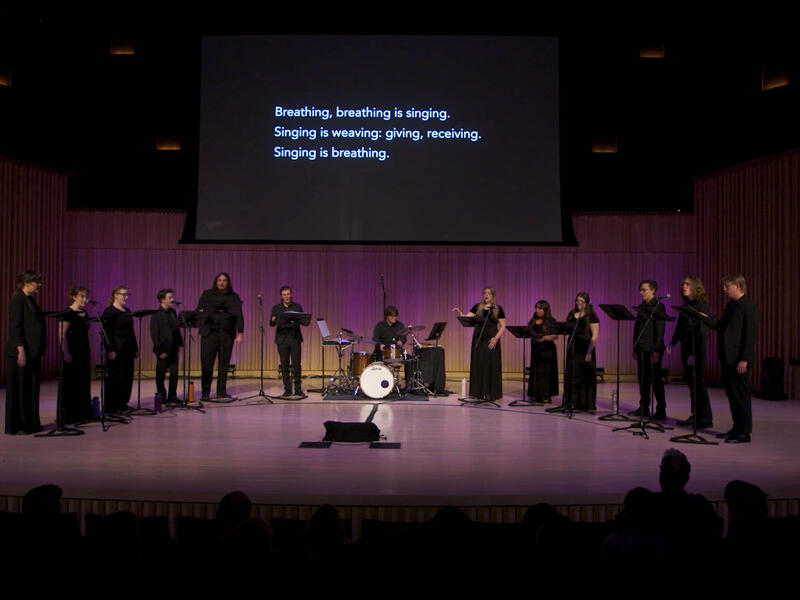 A photo of 11 singers on a stage with a drummer at a drum set sitting in the middle of the group. A black screen behind them has white text that reads \"Breathing, breathing is sining.\" \"Singing is giving: weaving and receiving.\" \"Singing is breathing.\"