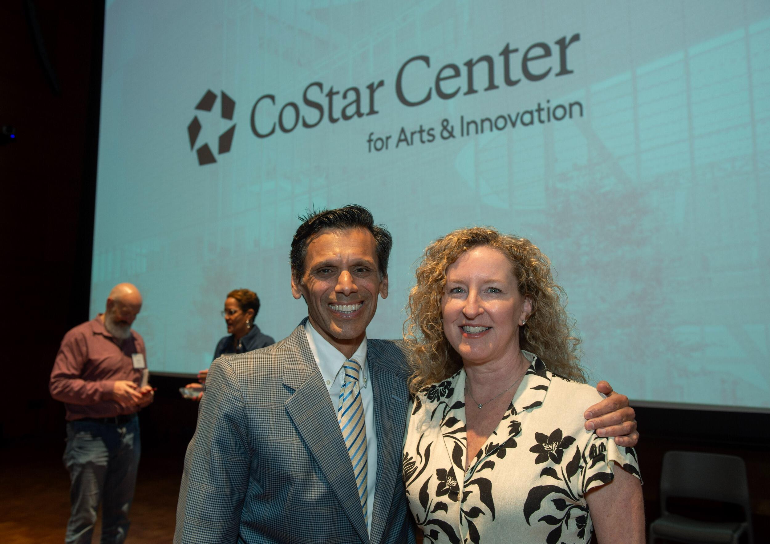 A man and a woman standing next to each other in front of a projected image that says \"CoStar Center for Arts and Innovation.\" The man is standing to the left of the woman and has his hand on her shoulder. 