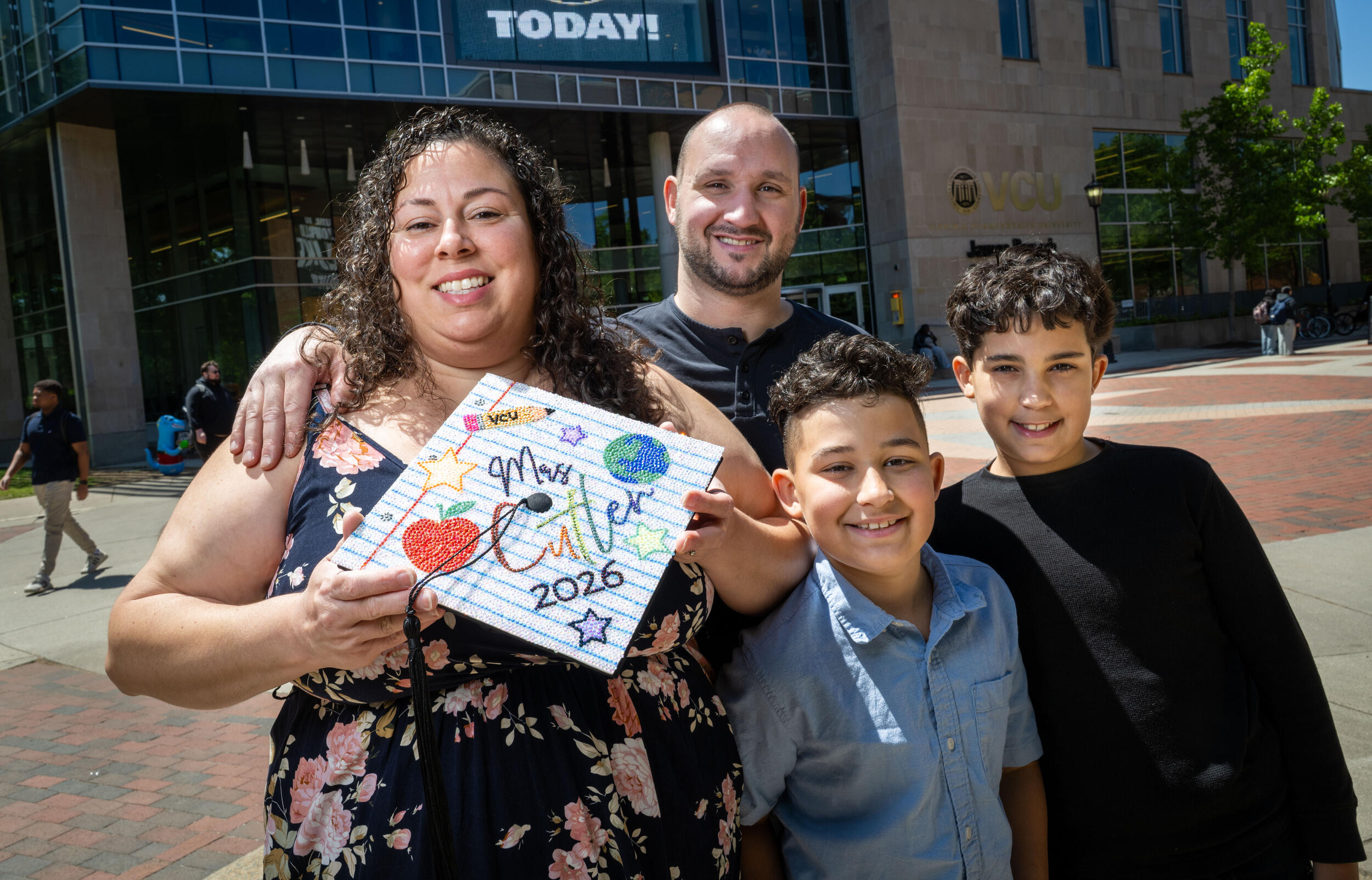 A photo of a woman, a man, and two boys. The woman is holding a graduation cap that is decorated to say \"Mrs. Cutler 2026.\"