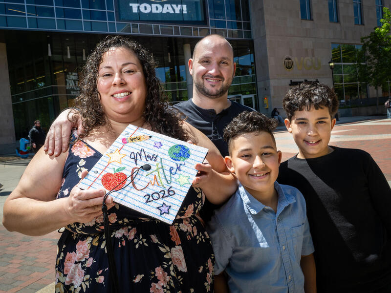 A photo of a woman, a man, and two boys. The woman is holding a graduation cap that is decorated to say \"Mrs. Cutler 2026.\"