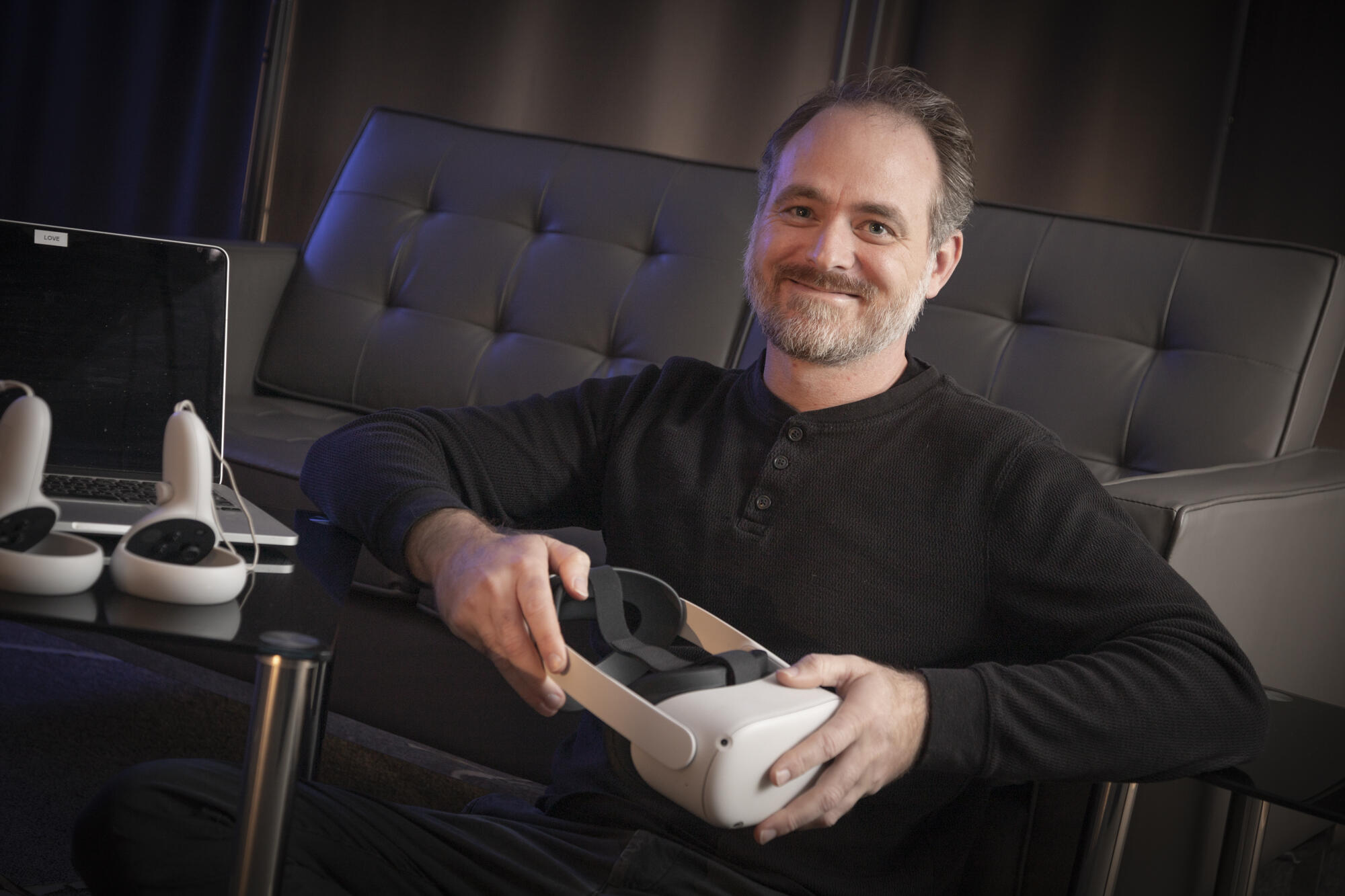 A photo of a man sitting by a couch and holding a VR headset. 