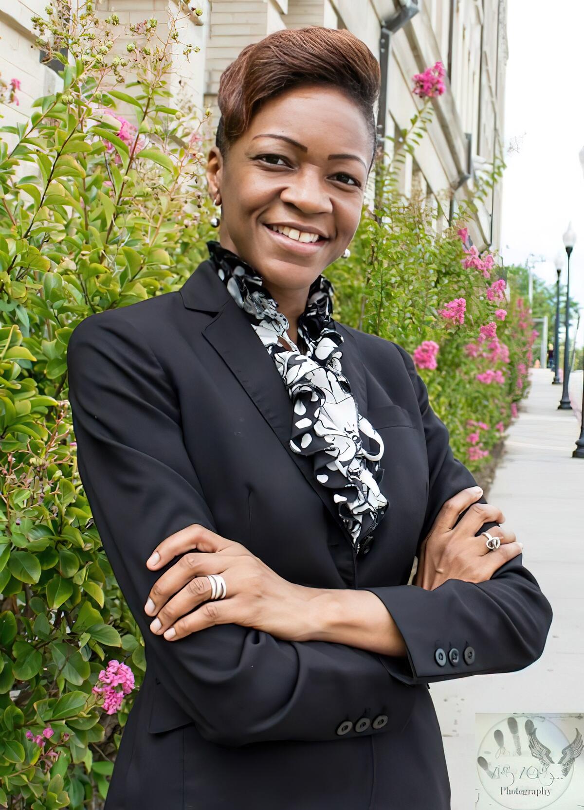 A photo of a woman from the waist up. She is standing with her arms crossed in front of her chest and is smiling. 